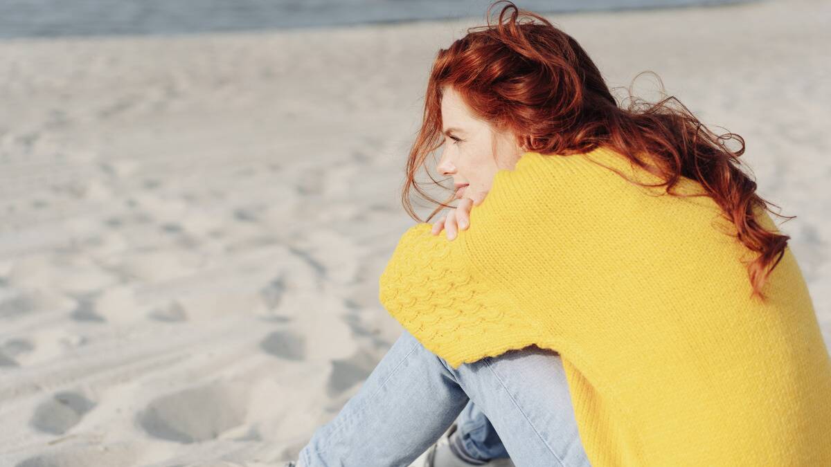 A woman sitting on the beach, knees drawn up to her chest, arms folded over them, chin resting on her arms.