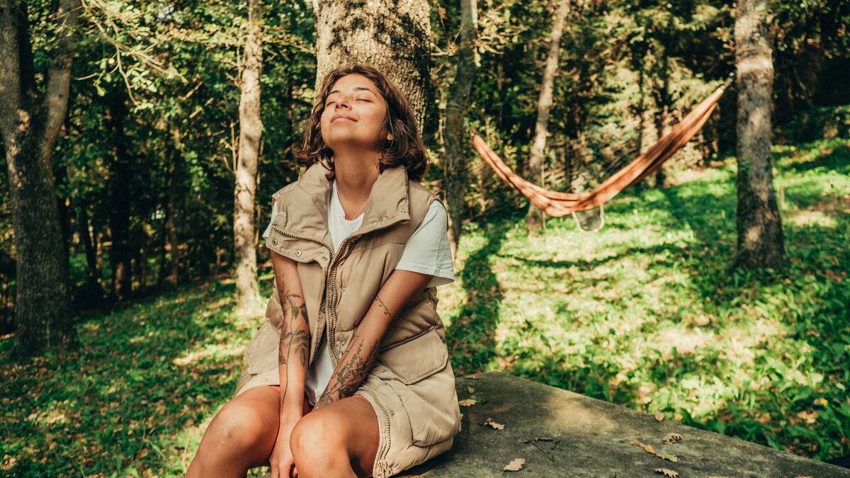 A woman sitting among the trees, eyes closed and smiling with her head tilted back.