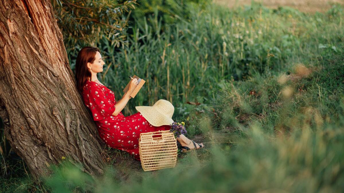 A woman in a bright red dress sitting at the base of a tree, writing in a notebook, smiling serenely.