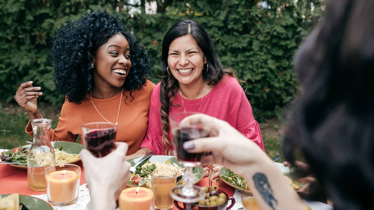 Two friends among a table full of people, sitting close and laughing as they chat.