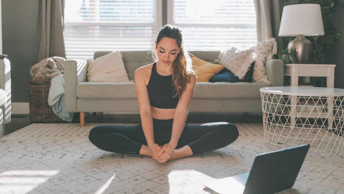 A woman doing yoga on her living room floor, in butterfly pose, her laptop open on the ground in front of her.