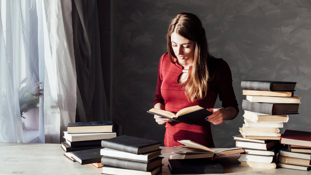 A woman standing at a table covered in books, looking down at an open one in her hands.