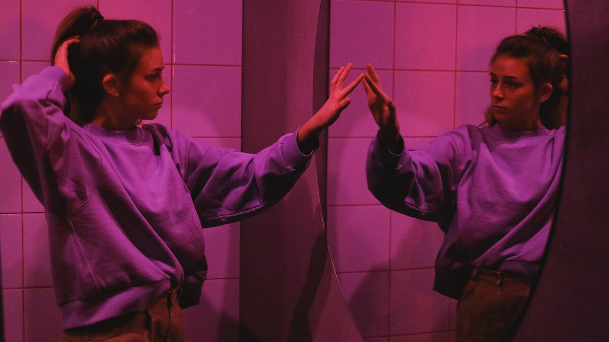 A woman in pink lighting looking at herself in a large mirror, reaching out to lightly touch her reflection.