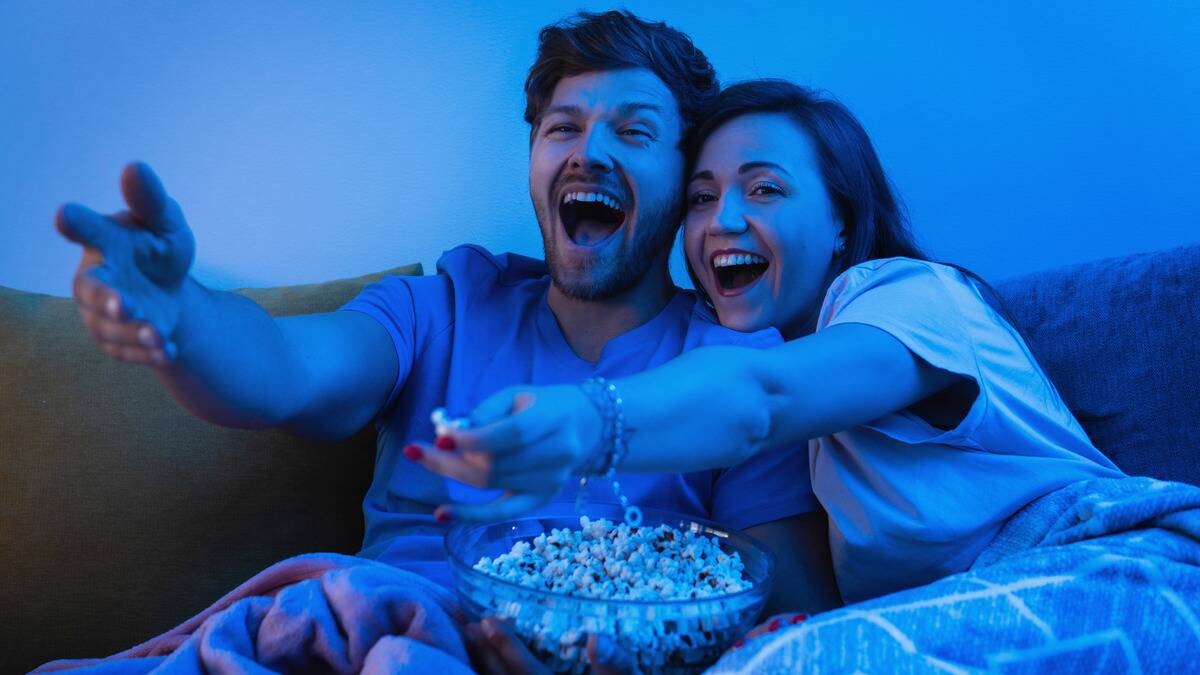 A couple cuddling on the couch while watching a movie, laughing and pointing at something on screen, a bowl of popcorn between them.