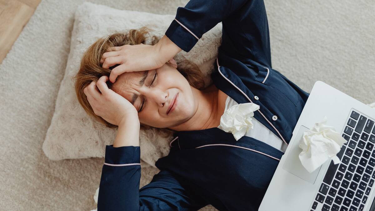 A woman laying down with her laptop on her stomach, hands on her head, looking stressed and upset, used tissues on her chest.