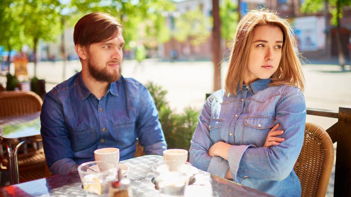 A woman and a man sitting at a cafe table, the man looking confused as the woman crosses her arms and turns her head away.