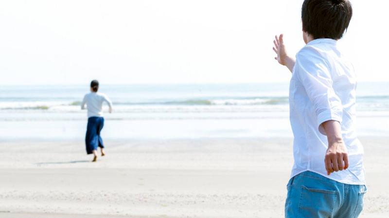 A woman in the distance running away from a man who's reaching out to her, both on the beach.