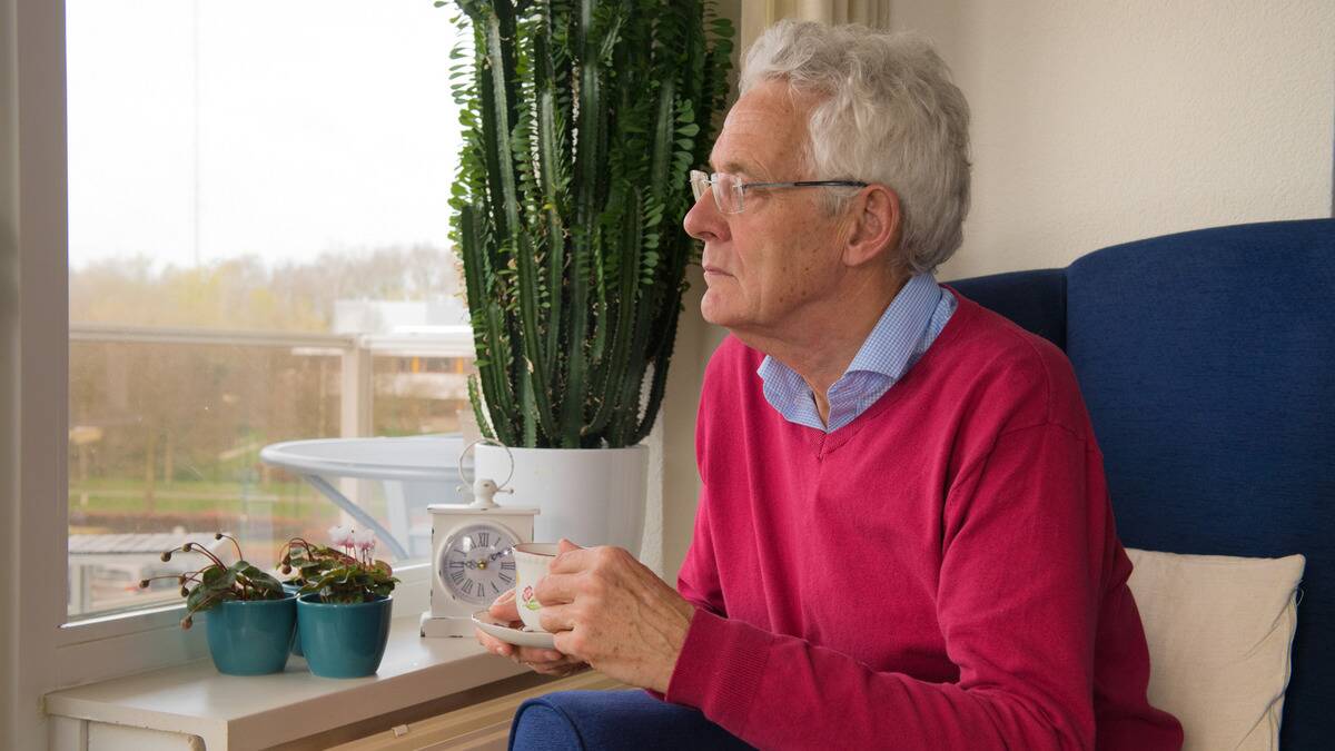 A man sitting in a chair holding a cup of tea, looking out the window forlornly.