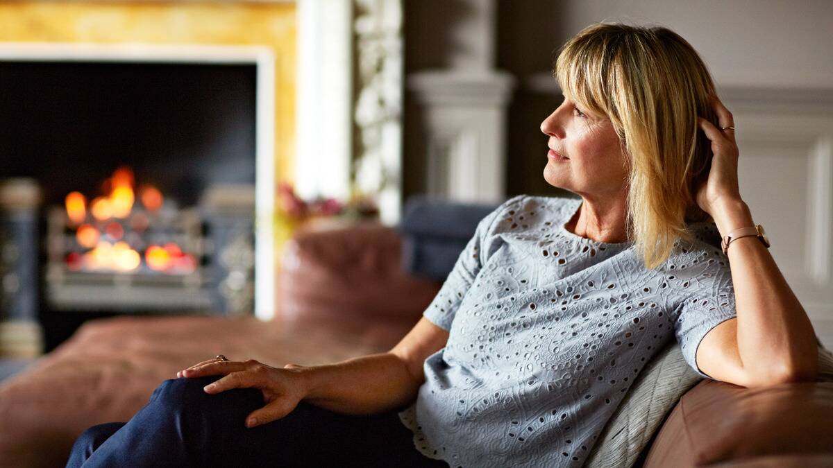 A woman sitting on a couch, leaning her head against her hand, smiling softly as she looks forward.