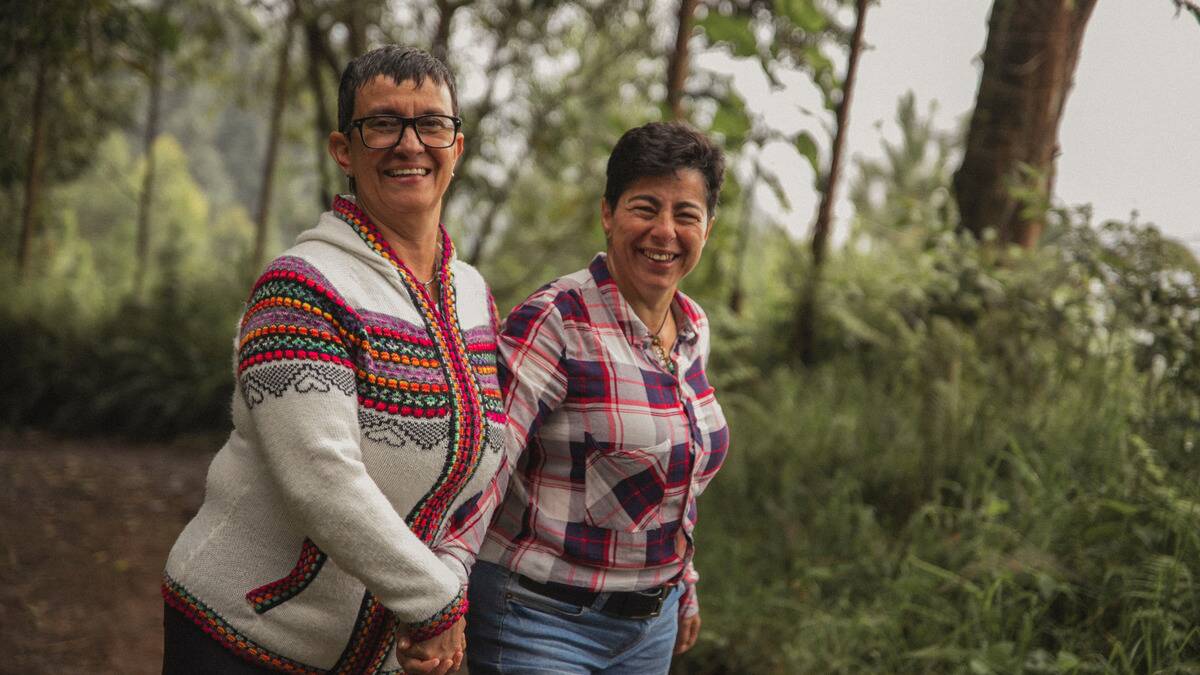 A couple holding hands and smiling at the camera as they walk a wooded path.