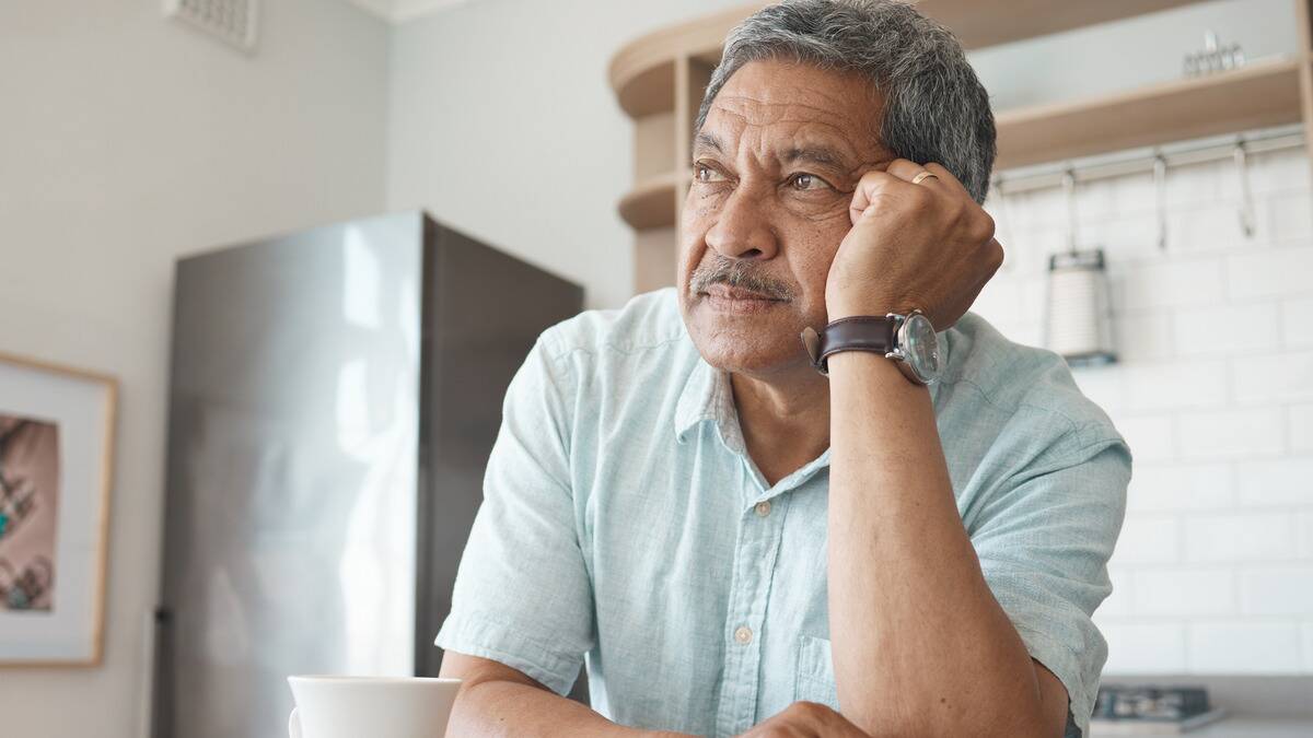 A man sitting at his kitchen counter, resting his face against his hand that's in a fist, looking into the distance thoughtfully.