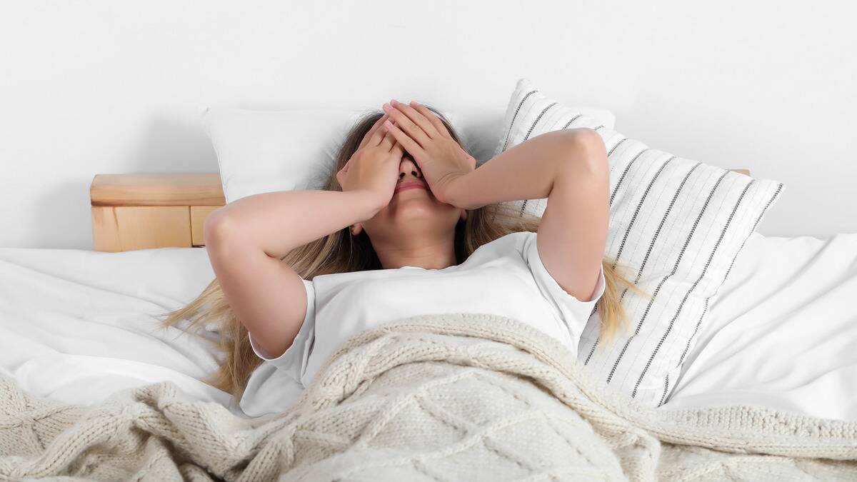 A woman laying on her back in bed, covering her face with both hands.