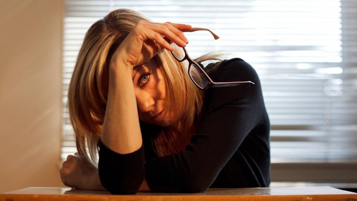 A woman sitting at a table, holding her glasses in her hand, half hiding her face behind her arm as she looks at the camera wearily.