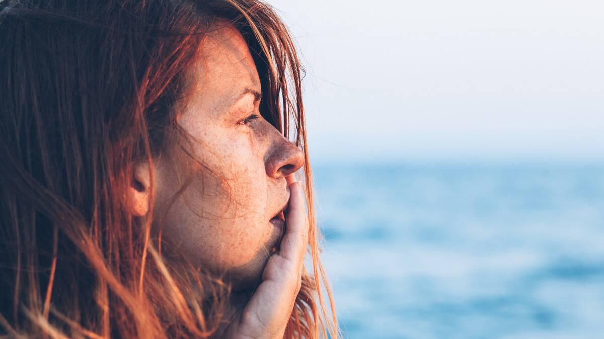 A close shot of a woman standing by the water, a hand on her cheek.