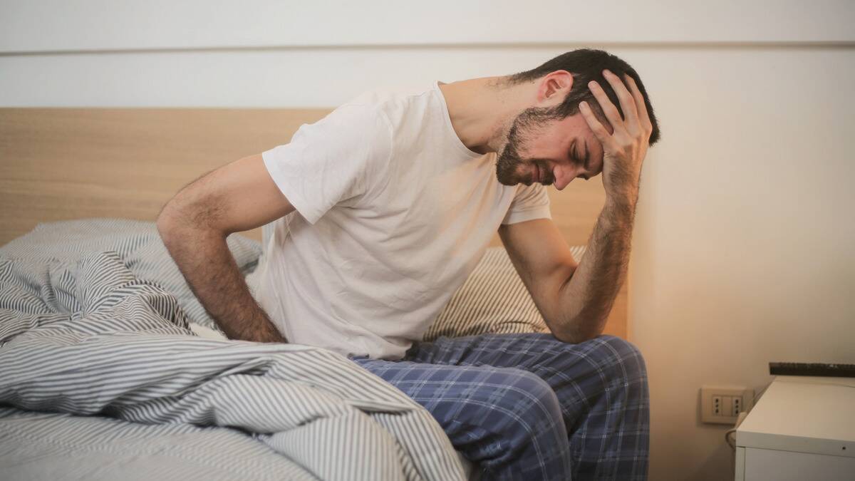 A man sitting up in bed, a hand on his forehead and another on his back, looking to be in pain.