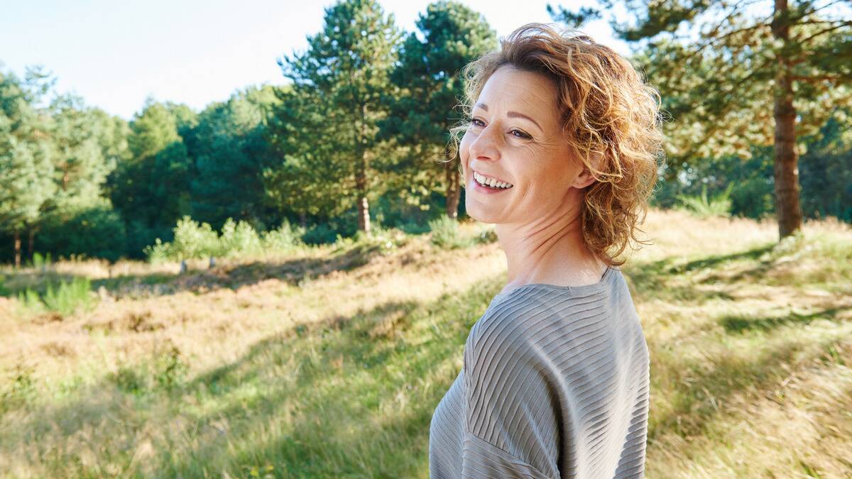 A woman standing outside in the sunshine, smiling as she looks over her shoulder.