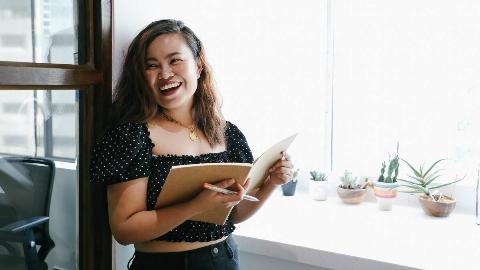 A woman smiling as she leans against a wall and holds a notebook open;