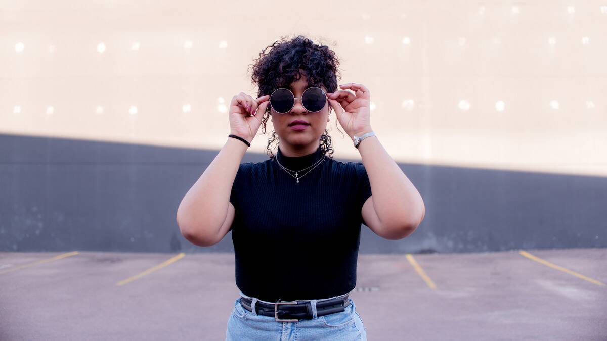 A stylish young woman standing in a parking lot looking directly at the camera, her hands holding the arms to her large, round sunglasses.