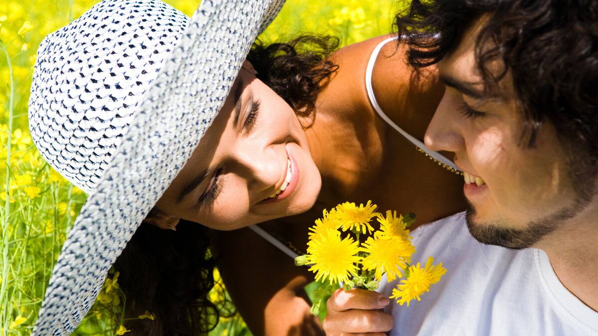 A couple sitting in the grass, the girl leaning over the man's shoulder, handing him a small bouquet of dandelions.