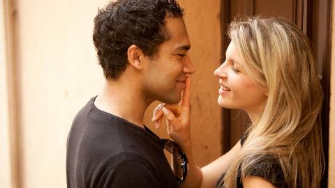 A couple standing close and flirting, the woman placing a finger over the man's lips.