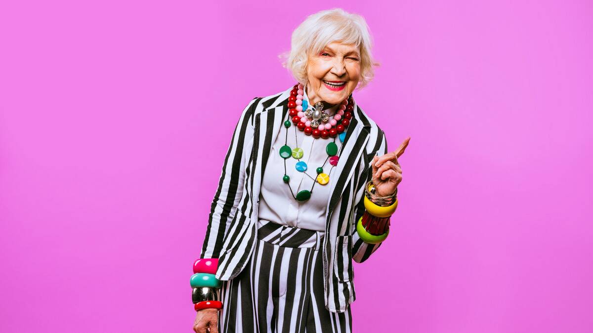 An older woman in a bold, unique outfit smiling cheekily in front of a bright pink backdrop.