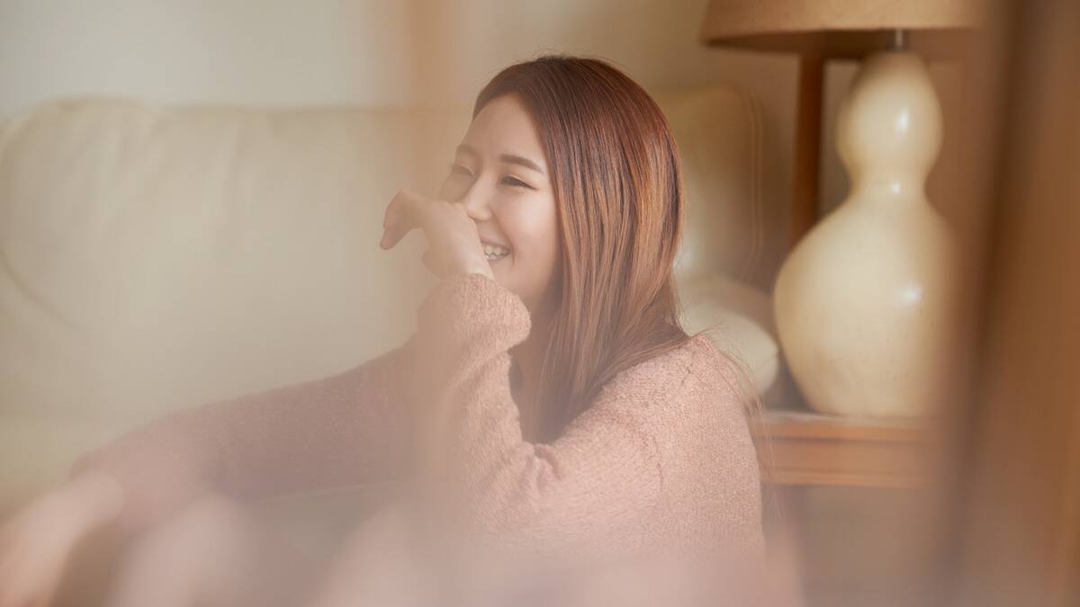 A woman smiling in her home, a hand raised to her mouth.