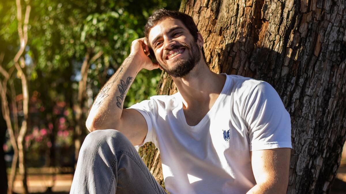 A man sitting at the base of a tree in the sunshine, smiling as he leans his head into his hand.