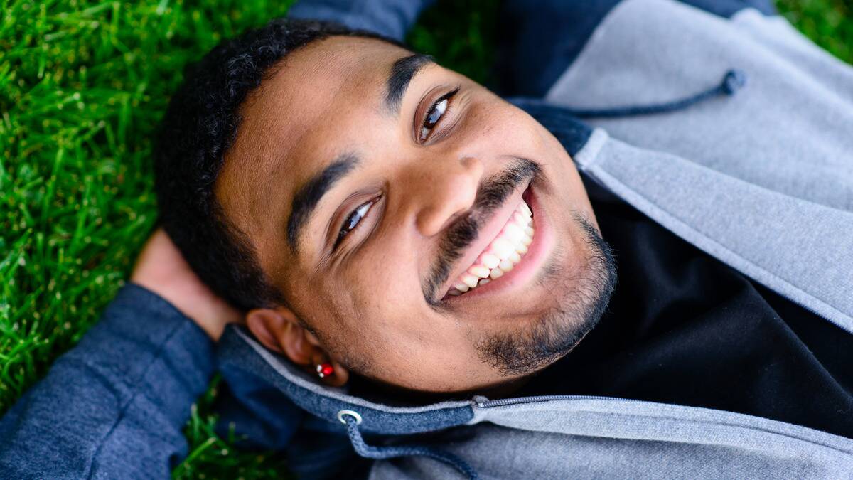 A close shot of a man smiling at the camera as he lays in the grass, hands behind his head.