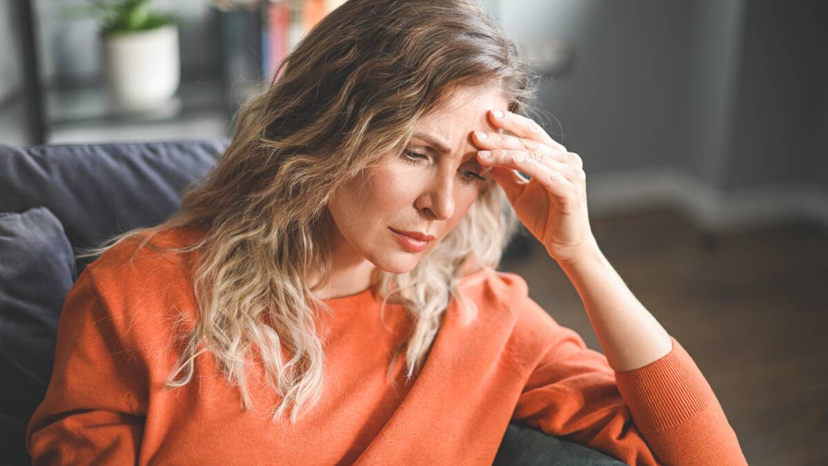 A woman sitting indoors, hand on her brow bone, looking concerned.