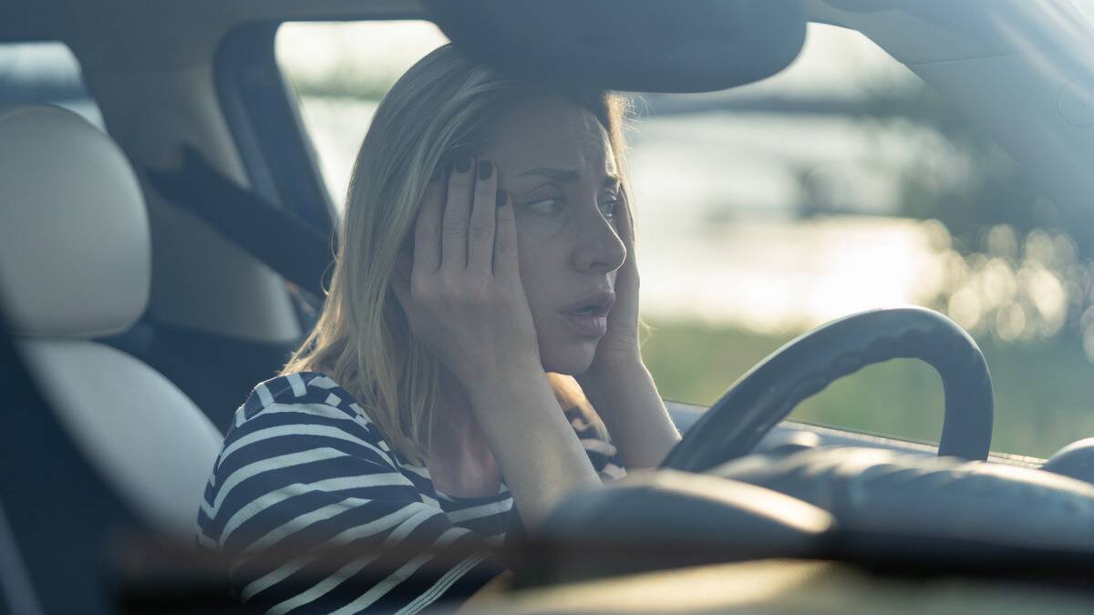 A woman sitting in her car, hands on both side of her face, looking worried.