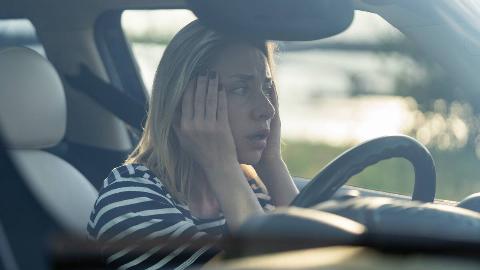 A woman sitting in her car, hands on both side of her face, looking worried.