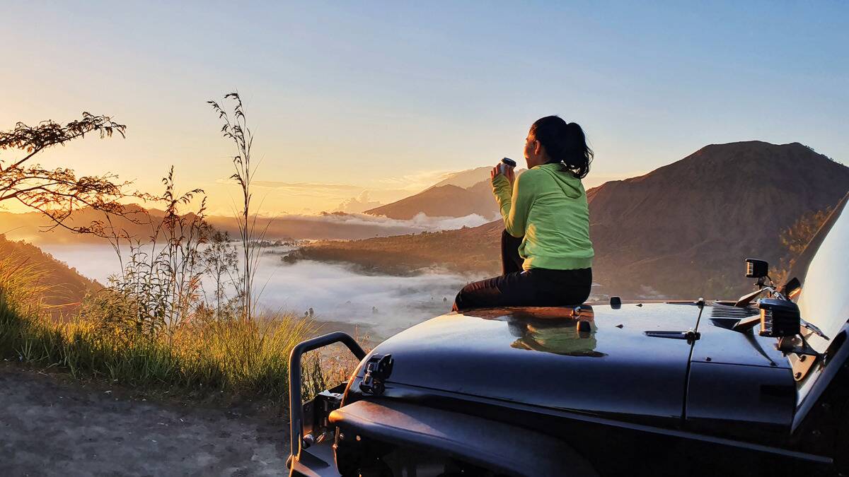 A woman sitting on the hood of her parked car, watching the sun rise by some mountains.