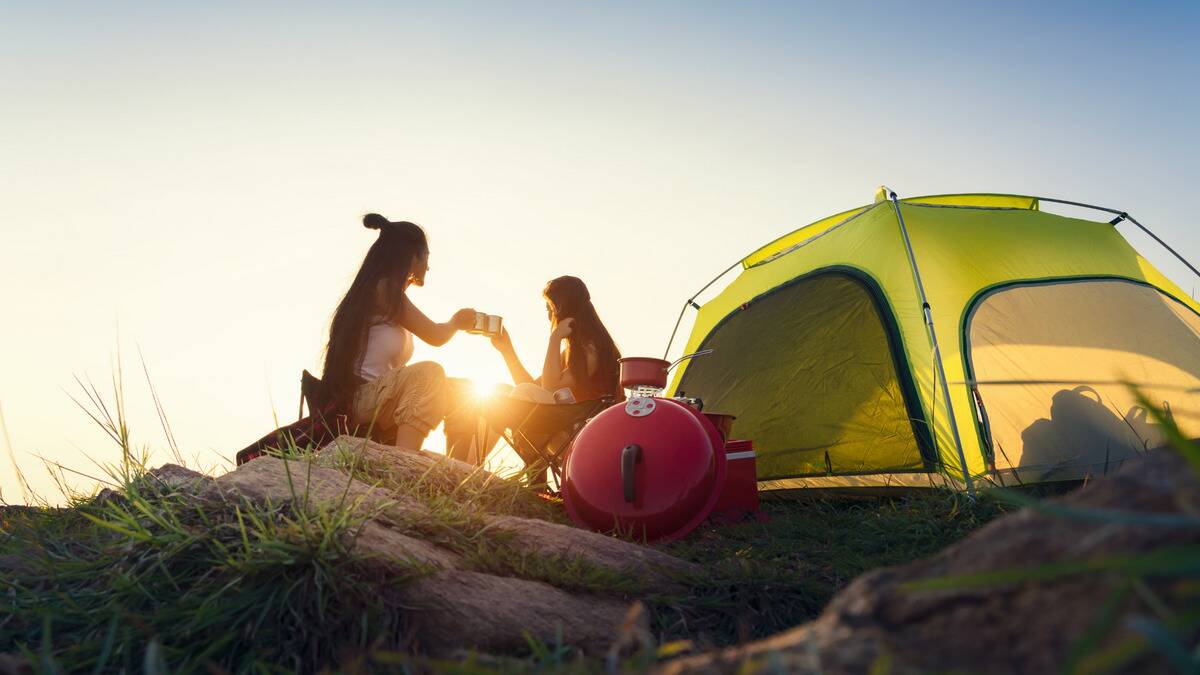 Two friends camping, the sun rising as they share mugs of coffee next to their tent.