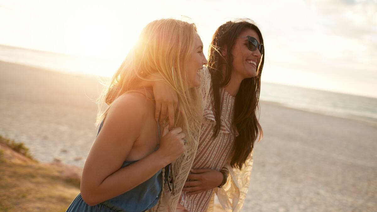 Two friends smiling and walking with their arms around one another as they walk down a beach.