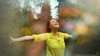 A woman standing out ina  forest, smiling at the sky with her arms out.