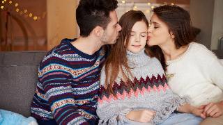 A daughter and her parents sitting on a couch, her eyes closed and smiling as her parents each kiss her on one cheek.