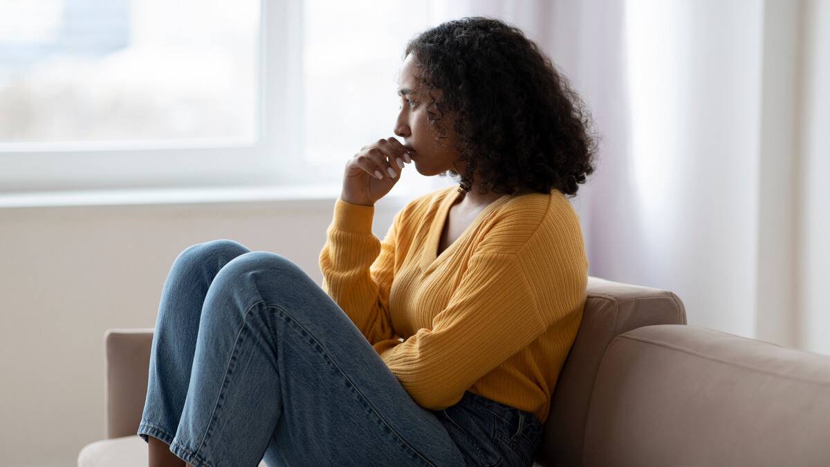 A woman sitting on a couch, knees drawn up, biting at her thumb nail.