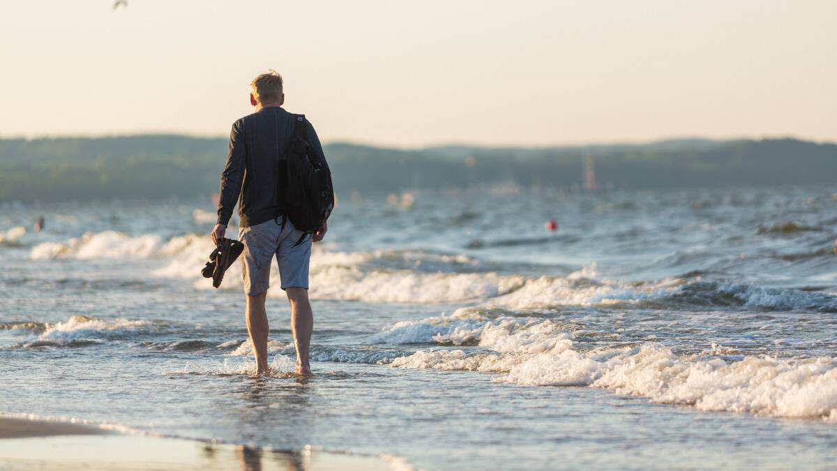 A man with his shoes in hand, standing ankle-high in wave-filled sea water.