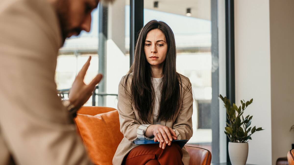 A woman sitting on a couch, legs and arms folded, looking insecure as a man speaks to her.