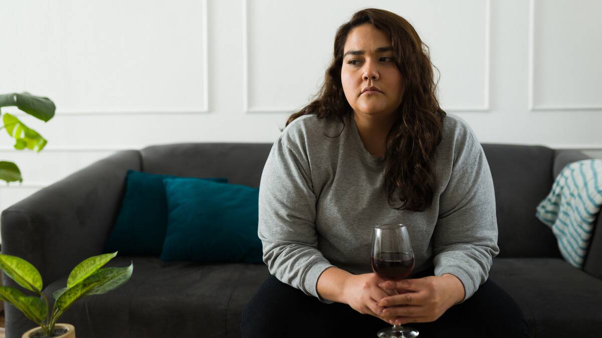 A woman sitting on he couch, holding a glass of wine, looking off to the side sadly.