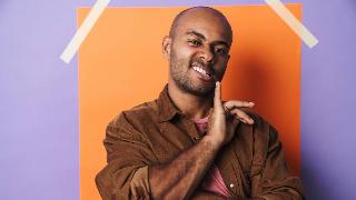 A man against a colorful background making a smarmy look at the camera, a finger lifted to his mouth.