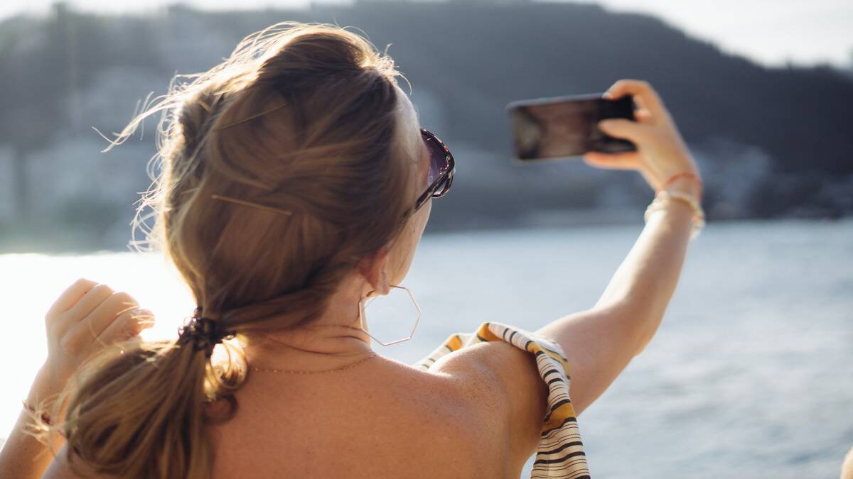 A woman sitting by a lake taking a selfie, seen over her shoulder.