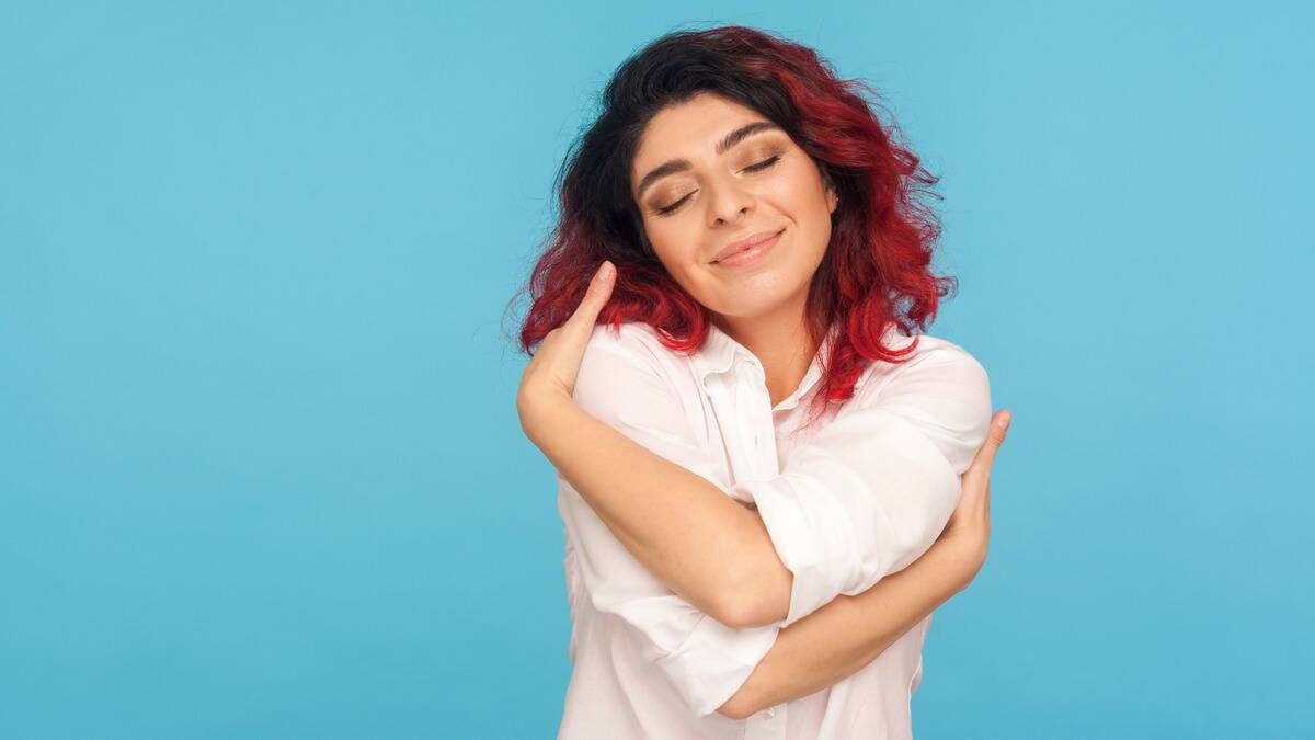 A woman against a bright blue background with her eyes closed, smiling as she hugs herself.