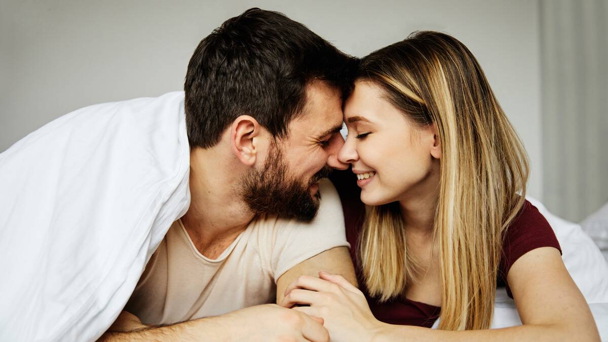 A couple laying on their stomachs in bed together, eyes closed, foreheads together, smiling.