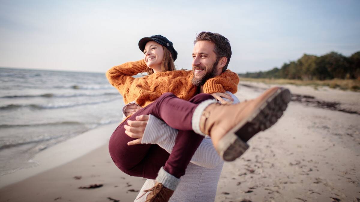 A man carrying his girlfriend bridal style along the beach, both smiling.