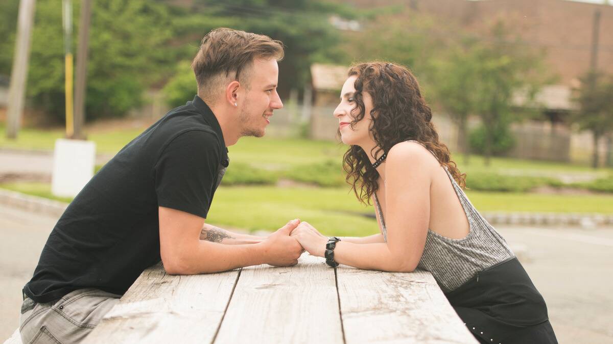 A couple sitting across each other at a picnic table, holding hands, leaning forward, smiling at each other.