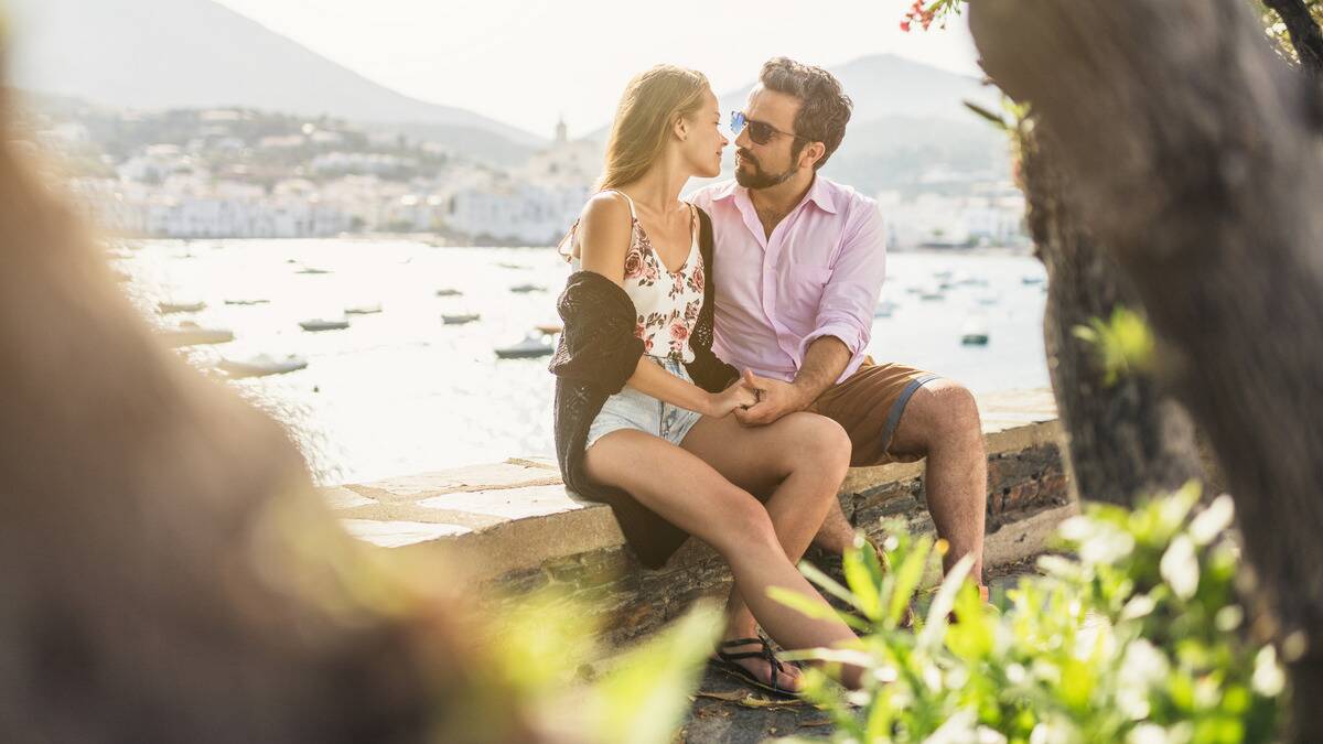 A couple sitting next to each other on a stone ledge by the water, seen through the branches of a tree.