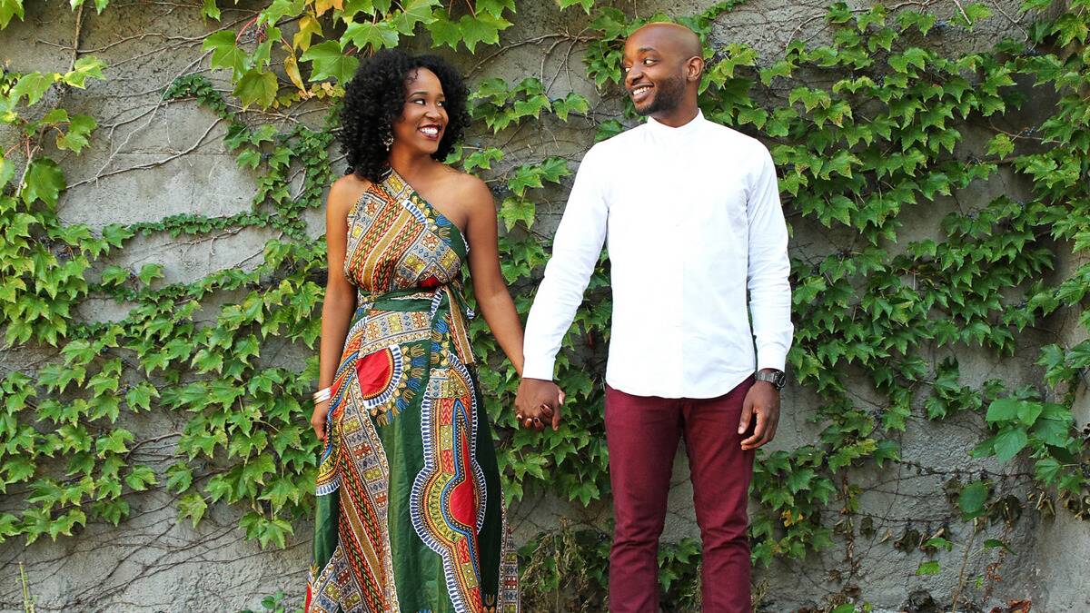 A couple standing side by side against an ivy-covered stone wall, holding hands and looking at each other with big smiles.