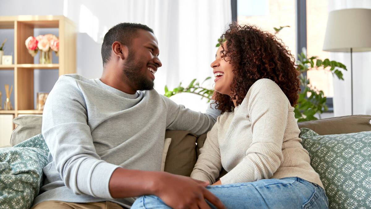 A couple sitting side by side on the couch, turned toward each other, laughing as they chat.