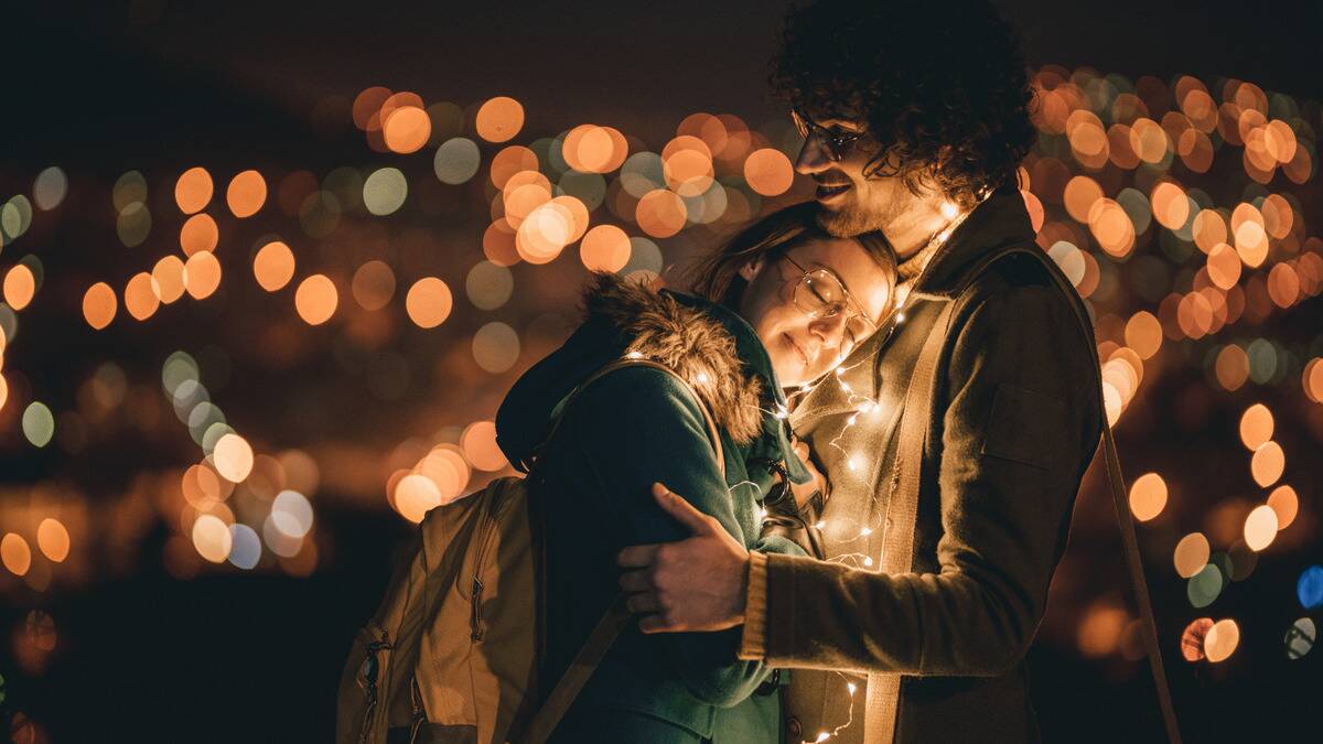 A couple hugging close outside at night, the woman leaning her head against the man's chest, a string of fairly lights around them.